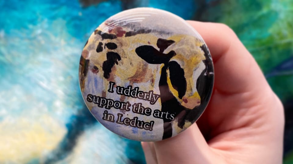 A colourful button with a cow saying "I udderly support the arts in Leduc!" being held up with an artistic, colourful background