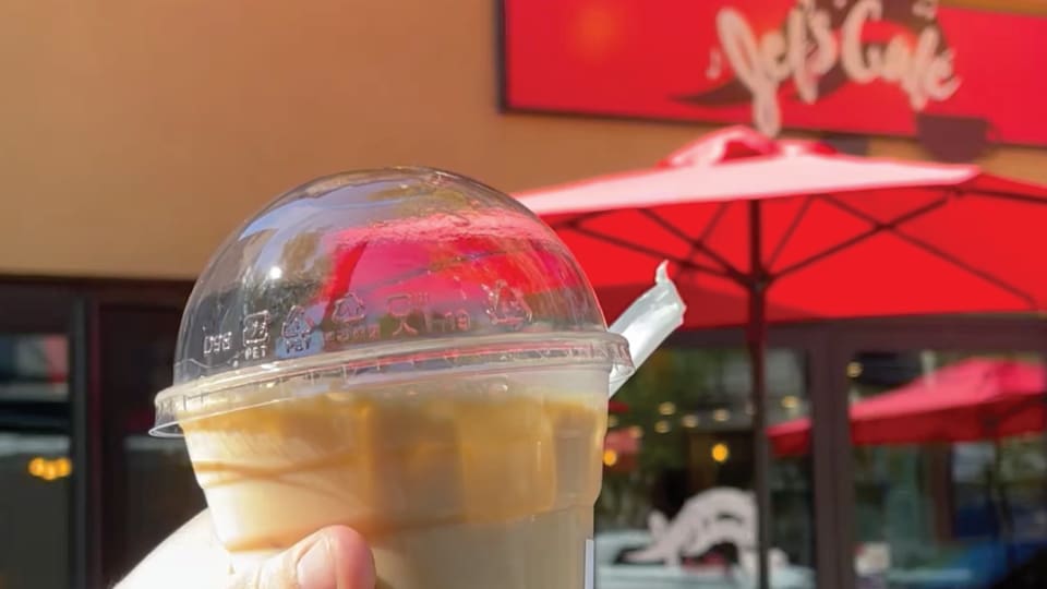 Holding an Iced coffee from the cafe outside in front of the sign and red patio umbrellas